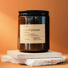 Load image into Gallery viewer, Candle jar labeled 'pride &amp; prejudice' on a stone surface with an orange background
