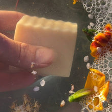 Load image into Gallery viewer, Bar of soap held by a hand with a soapy water background and flowers.
