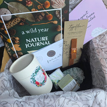 Load image into Gallery viewer, A curated gift box filled with various items including a 'a wild year NATURE JOURNAL', Bath Tea, quote cards, a brown leather accessory, and other small packaged goods, all arranged in patterned tissue paper inside a cardboard box.
