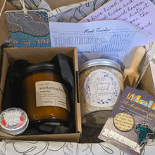 Load image into Gallery viewer, A curated gift box filled with self-care and wellness items, including a candle, a jar of bath salts, a book tracker card, a small wooden spoon, and a pair of bookish earrings, arranged in a cardboard box on patterned paper.
